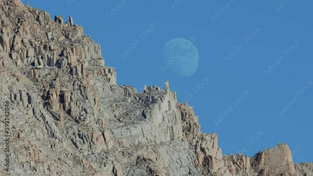 Large moon sits right behind rugged rocky mountain with a deep blue sky during the day (60 fps slowed to 30 fps) Large moon sits right behind rugged rocky mountain with a deep blue sky during the day (60 fps slowed to 30 fps)