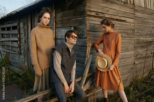 Portrait of three young beautiful people in village area. Wooden farm. Backyard. Nature. Yellow grass. Sunset light, autumn. Country style clothes