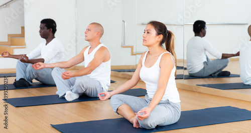 Focused women and men sitting on mats in fitness center, making yoga meditation in lotus pose .