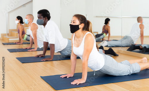 Adult women and men wearing protective masks performing yoga exercises during group workout in gym. Healthy lifestyle and pandemic precautions concept.