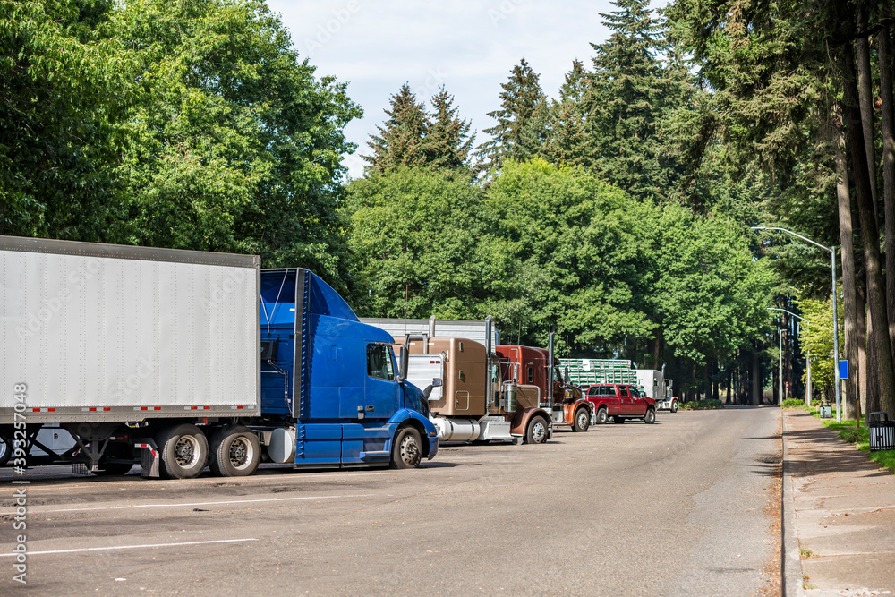 Line of different big rigs semi trucks and semi trailers standing on the forest rest area parking lot for take a brake