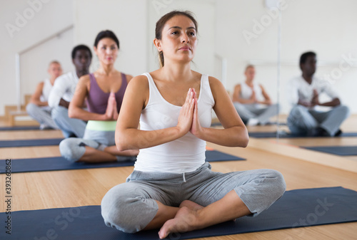 Portrait of young woman sitting in lotus position practicing meditation during group yoga class