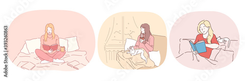 Relaxation with book at home, lazy bedding time concept. Young positive women in comfortable home clothing staying in bed, reading books and relaxing with cat and hot drink at home 