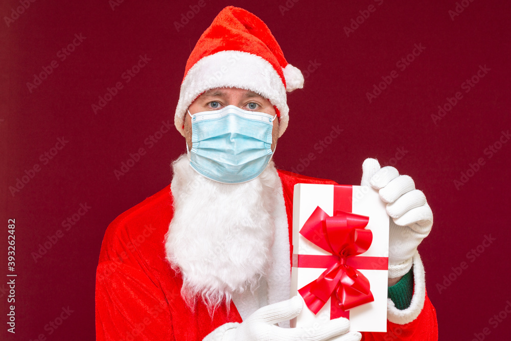 Christmas gift. Santa Claus wears medical mask holding gift box and looking at camera. red studio background