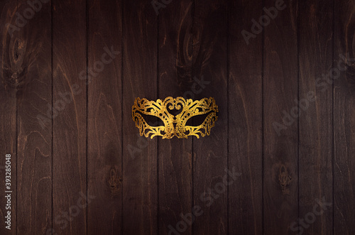 Golden glittering lace ornate theatre mask for masquerade on dark wood board, top view.