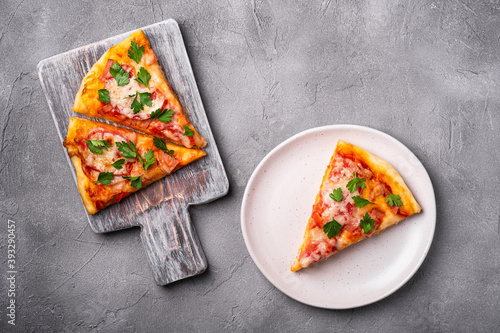 Hot pizza slices with mozzarella cheese, ham, tomato and parsley on wooden cutting board and plate, stone concrete background, top view