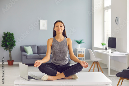 Relaxed slender asian girl sitting in lotus position on desk near laptop in office and meditating.
