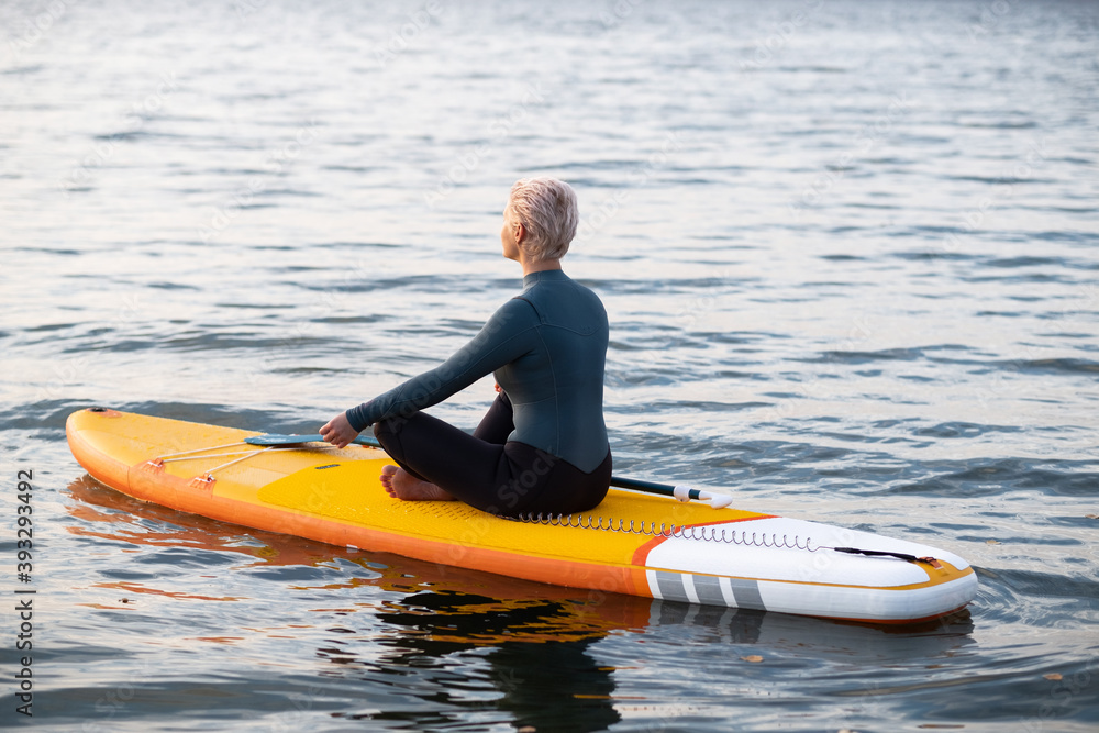 Obraz premium A young athletic woman in a blue wetsuit swims through the sea on a yellow paddle Board and meditates in the Golden rays of the dawn sun