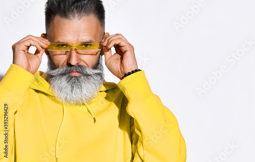 Portrait of serious brutal bearded middle aged man in yellow hoodie standing and putting on glasses over white wall background. Stylish bright comfortable casul fashion for men concept photo