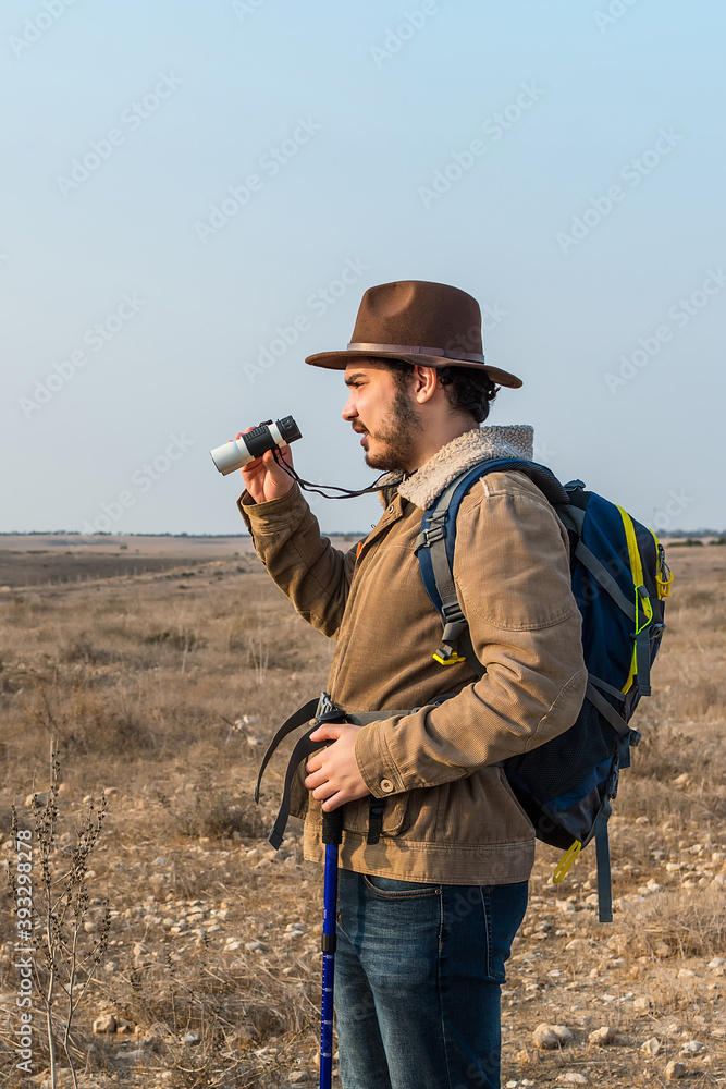 Hiker young man with beard, wearing brown jacket, backpack and a hipster hat look through binoculars. Hiker young man with beard, wearing brown jacket, backpack and a hipster hat look through binoculars.