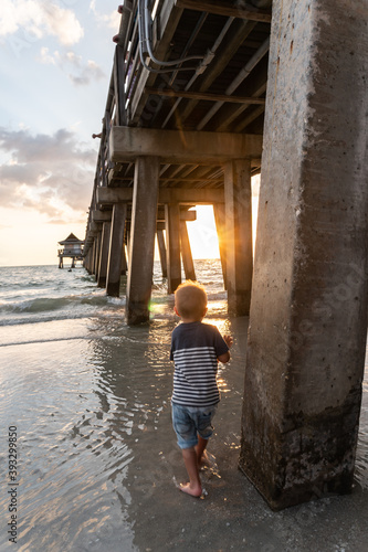 Boy standing under the pier in water watching sunset through concrete supports