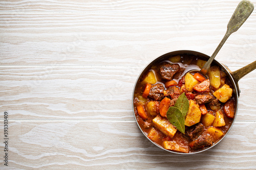 Delicious stew with meat, potatoes, carrot and gravy in rustic copper pot on white wooden background from above. Traditional winter and autumn dish beef and vegetables ragout in stewpot, copy space
