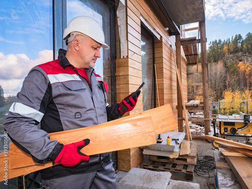 Builder with wooden boards in his hands. Builder is calling someone. Concept - he orders lumber by phone. Purchase of lumber. A man in a builder's uniform. He outside a house under construction. © Grispb