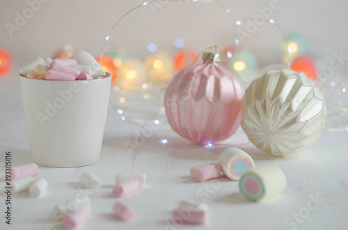 White cup with marshmallows and Christmas balls on a blurred background.