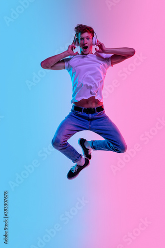 Music in headphones. Young caucasian man's jumping high on gradient blue-pink studio background in neon light. Concept of youth, human emotions, facial expression, sales, ad. Beautiful model in casual