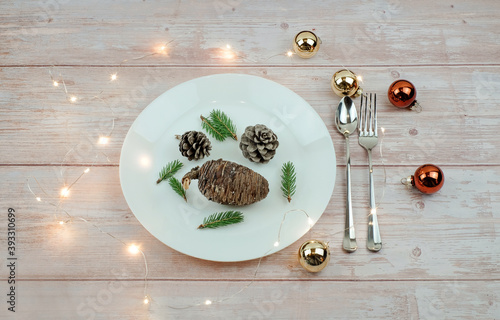 Christmas diet concept. Pine cones and pine branches as food on whete plate, fork and knife on wooden table, lights and balls decoration. Vegan menu concept. photo