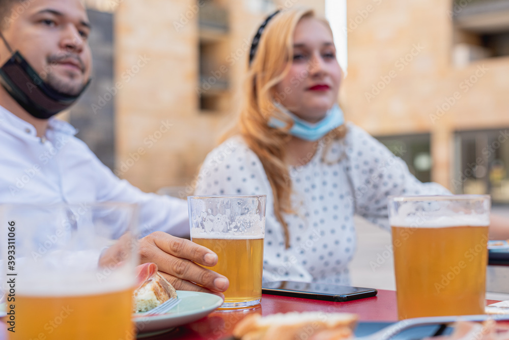 glasses of beer at the table during hangout of a millennial group of people, young couple chat at the table on the blurred background using facemask in a incorrect way, the new normal socialization glasses of beer at the table during hangout of a millennial group of people, young couple chat at the table on the blurred background using facemask in a incorrect way, the new normal socialization