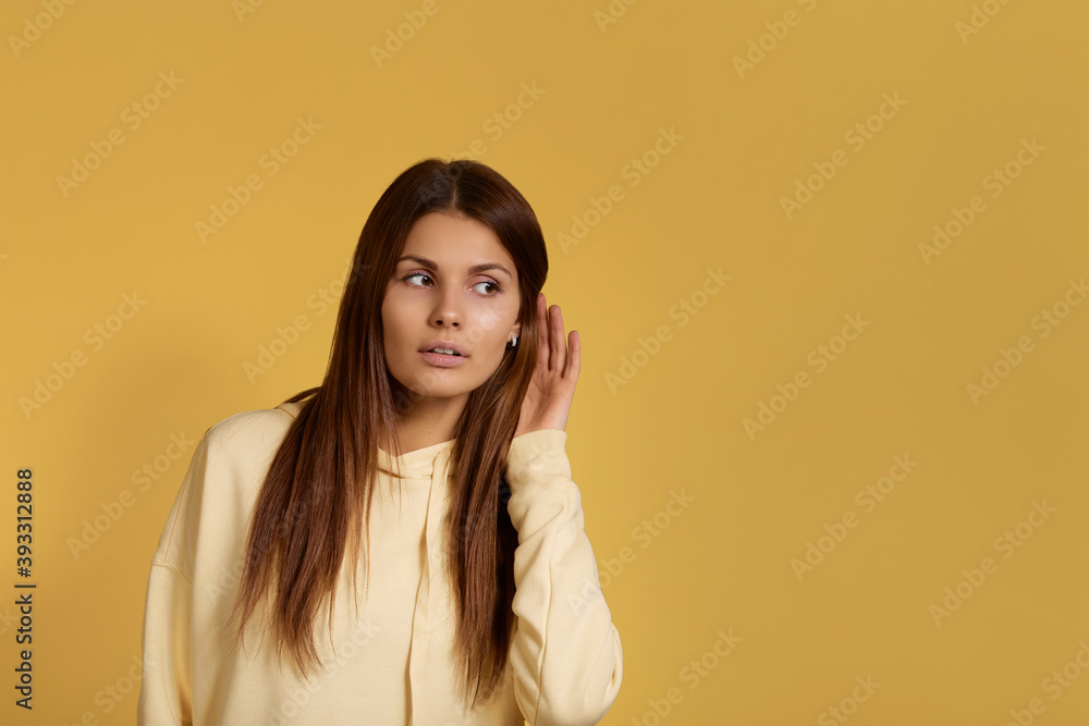 Like a spy. Impressed caucasian woman in yellow hoodie tries to hear details of rumor, holds hand near ear, eavesdrops with great interest. Isolated over a yellow background. Like a spy. Impressed caucasian woman in yellow hoodie tries to hear details of rumor, holds hand near ear, eavesdrops with great interest. Isolated over a yellow background.