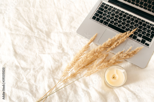 Laptop, beige reeds agains pampas plumen, candle on linen white bed. Minimal, stylish, trend concept. Autumn, fall, composition. Flat lay, top view, copy space. photo