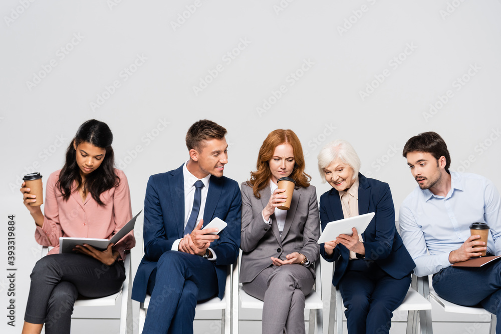 Senior businesswoman showing digital tablet near multiethnic businesspeople with coffee to go isolated on grey Senior businesswoman showing digital tablet near multiethnic businesspeople with coffee to go isolated on grey