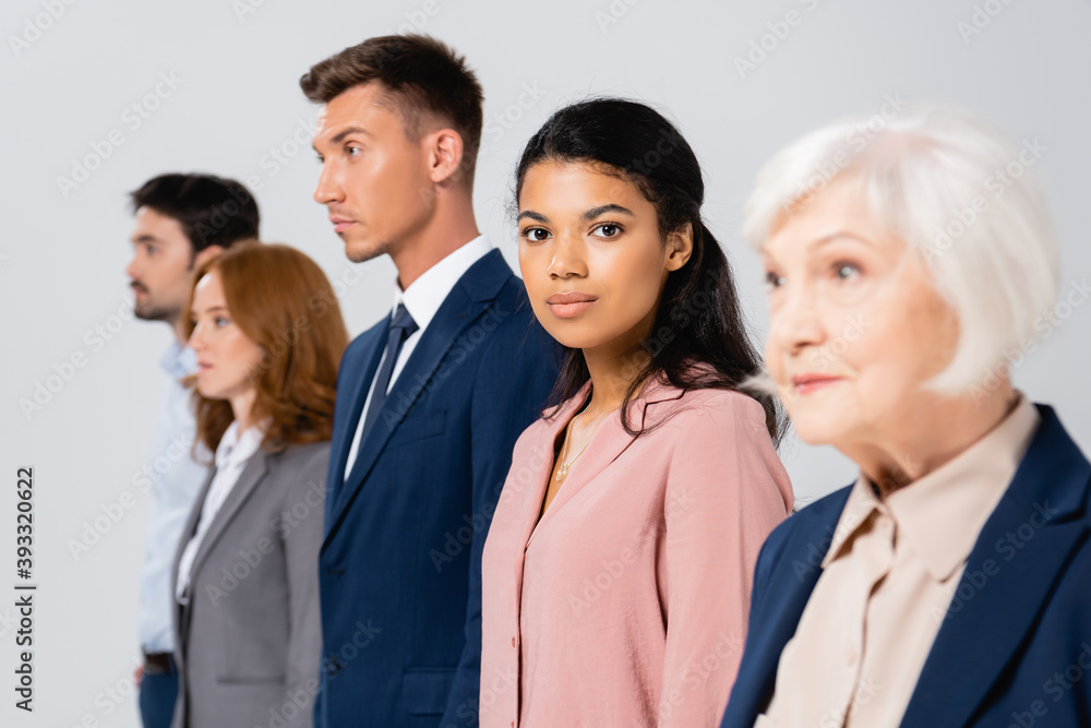 African american businesswoman looking at camera near colleagues on blurred background isolated on grey African american businesswoman looking at camera near colleagues on blurred background isolated on grey