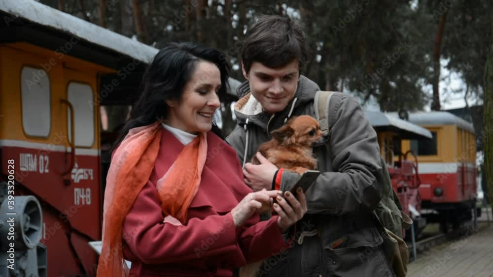 A woman and a young guy with a dog are watching something on the phone in a city park A woman and a young guy with a dog are watching something on the phone in a city park