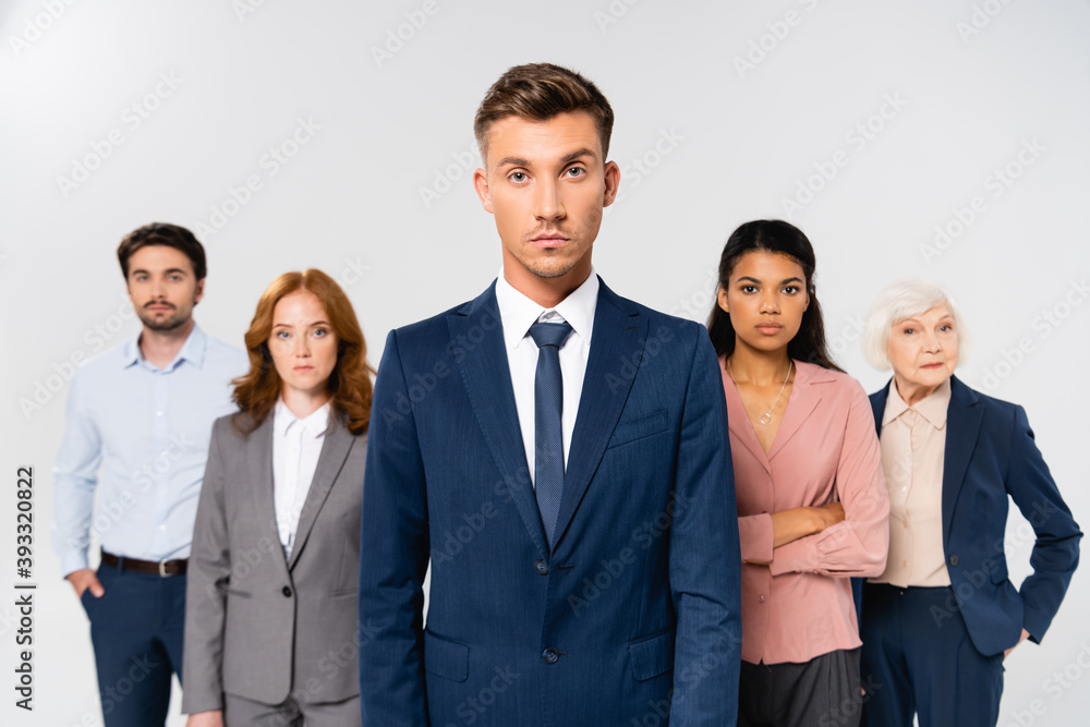 Businessman in suit looking at camera near multicultural colleagues on blurred background isolated on grey Businessman in suit looking at camera near multicultural colleagues on blurred background isolated on grey