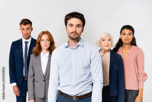 Young businessman looking at camera while standing near multicultural businesspeople on blurred background isolated on grey