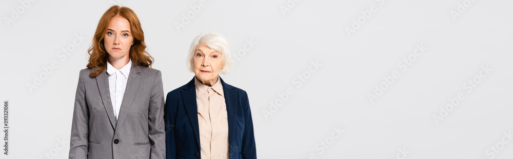 Red haired and senior businesswomen looking at camera isolated on grey, banner Red haired and senior businesswomen looking at camera isolated on grey, banner