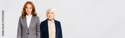 Red haired and senior businesswomen looking at camera isolated on grey, banner