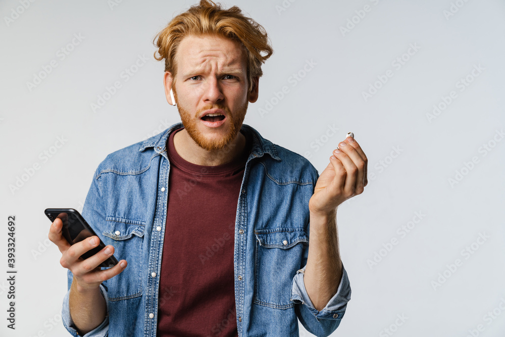 Confused redhead guy in wireless earphones using cellphone Confused redhead guy in wireless earphones using cellphone
