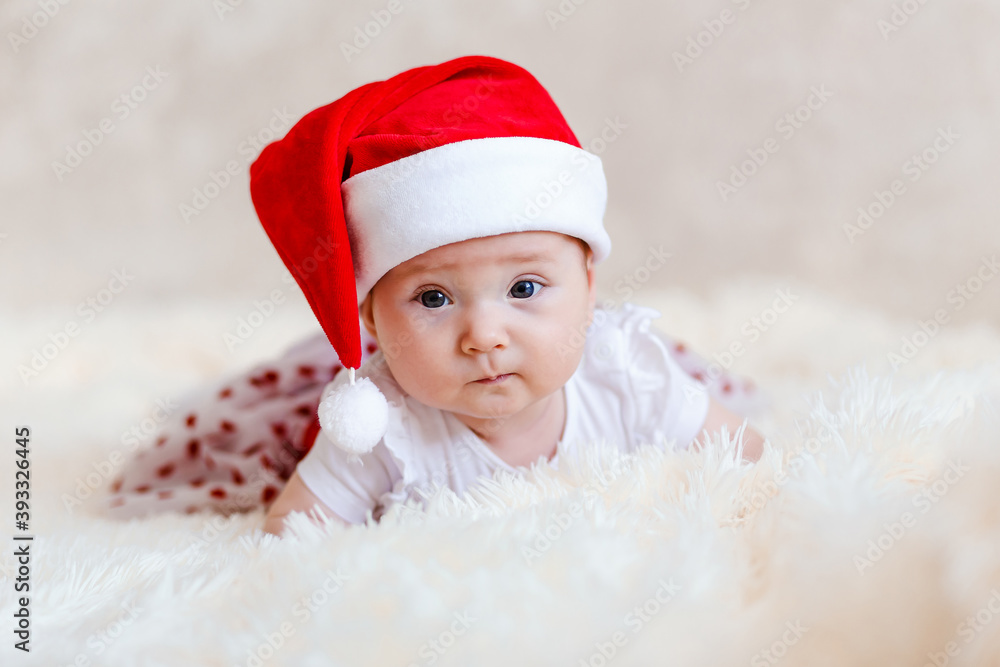 European six-month-old baby girl with big beautiful eyes in red santa cap lies on her stomach for Christmas. Photo shoot babies European six-month-old baby girl with big beautiful eyes in red santa cap lies on her stomach for Christmas. Photo shoot babies