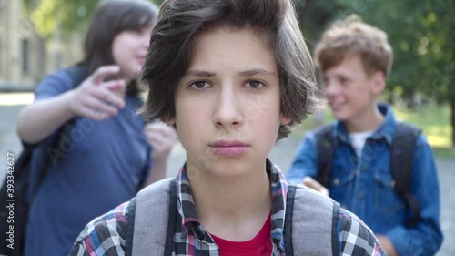 Close-up of bullied brunette schoolboy with brown eyes looking at camera as blurred classmates pointing at him and laughing at the background. Sad bullying victim posing outdoors.