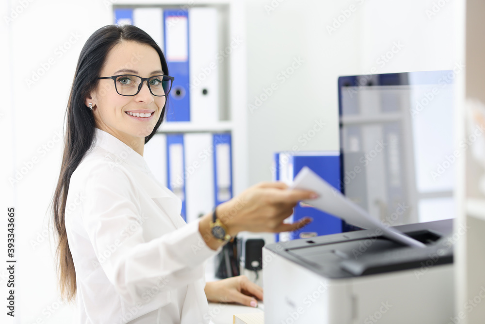 Smiling portrait of secretary who is holding papers next to the printer. Office vacancies without work experience concept Smiling portrait of secretary who is holding papers next to the printer. Office vacancies without work experience concept
