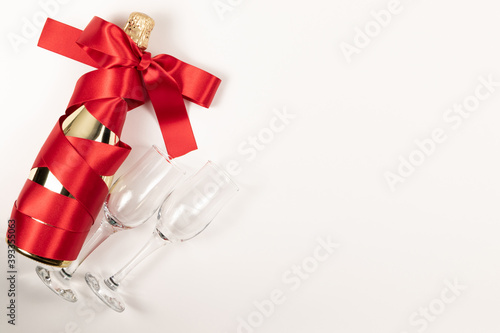 gift bottle of champagne in gold foil wrapped in a red silk ribbon and two glasses on a white background