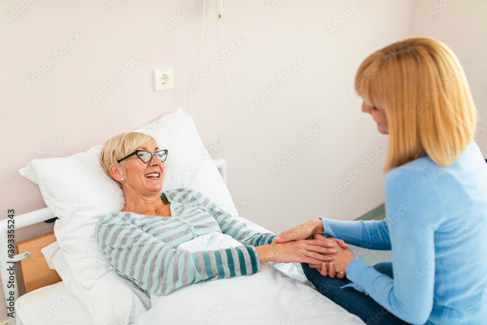 Fototapeta premium Beautiful middle age woman spending some time with her mother in hospital or nursing center. Senior woman lying in bed.