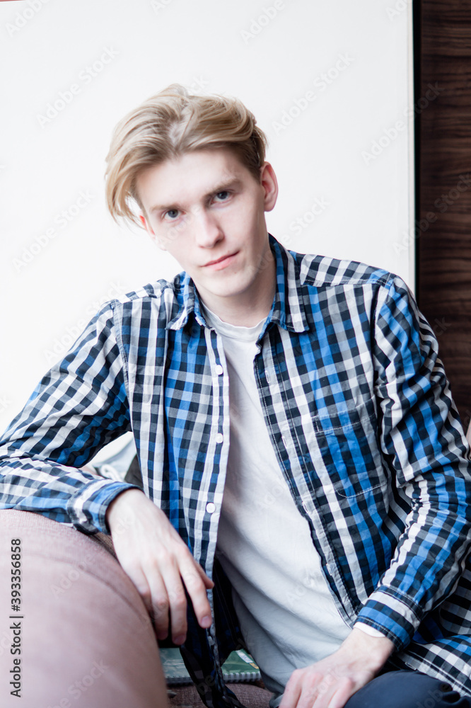 a young guy in a checkered shirt sits on the couch, looks at the camera, long hair on his head, is waiting for a meeting a young guy in a checkered shirt sits on the couch, looks at the camera, long hair on his head, is waiting for a meeting