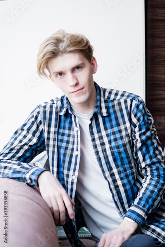a young guy in a checkered shirt sits on the couch, looks at the camera, long hair on his head, is waiting for a meeting © Maxim