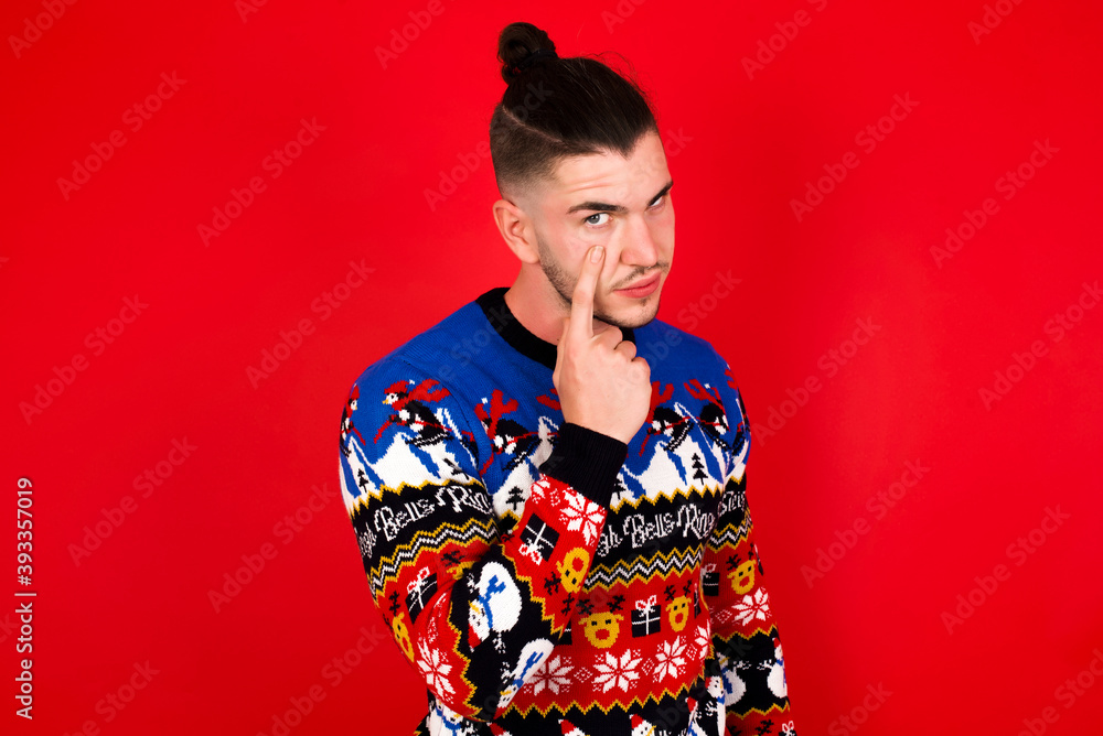 Young handsome Caucasian man wearing Christmas sweater against red wall, Pointing to the eye watching you gesture, suspicious expression. Young handsome Caucasian man wearing Christmas sweater against red wall, Pointing to the eye watching you gesture, suspicious expression.