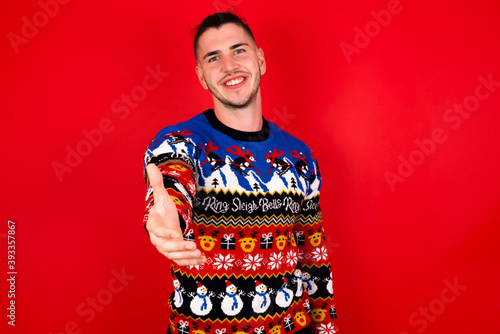 Young handsome Caucasian man wearing Christmas sweater against red wall, smiling friendly offering handshake as greeting and welcoming. Successful business.