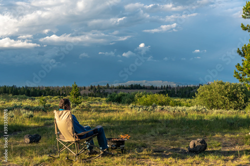 A man sitting on a rocking chair enjoying a campfire and the view of distance mountain under dramatic clouds illuminated by the setting sun, Baker's Hole Campground, West Yellowstone, Montana