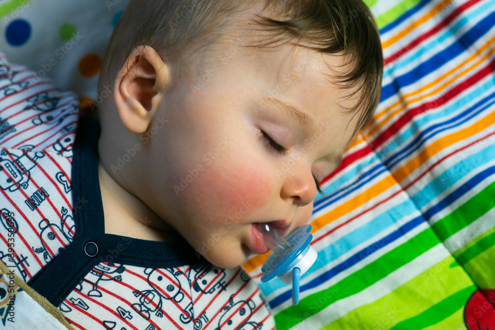 Little caucasian baby boy with red cheeks sleeping, holding his pacifier in mouth on colorful blanket background.