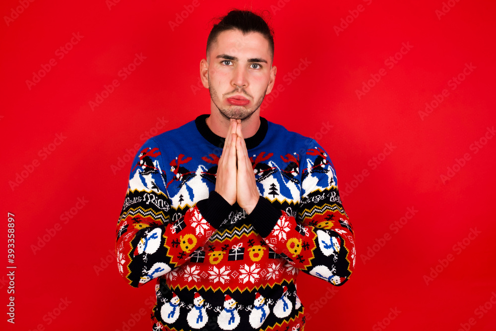Positive Young handsome Caucasian man wearing Christmas sweater against red wall,  smiles happily, glad to receive pleasant news from interlocutor, keeps palms together, People emotions concept.