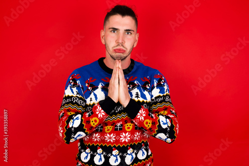 Positive Young handsome Caucasian man wearing Christmas sweater against red wall, smiles happily, glad to receive pleasant news from interlocutor, keeps palms together, People emotions concept.