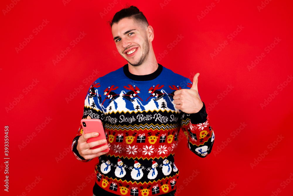 Portrait of Young handsome Caucasian man wearing Christmas sweater against red wall, using and texting with smartphone happy with big smile doing ok sign, thumb up with fingers, excellent sign Portrait of Young handsome Caucasian man wearing Christmas sweater against red wall, using and texting with smartphone happy with big smile doing ok sign, thumb up with fingers, excellent sign