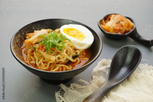 Instan ramyeon or Korean noodles soup on black bowl, topped with half boiled egg, leek and Kimchi on small bowl