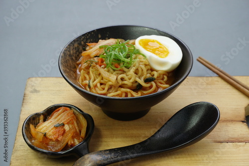 Instan ramyeon or Korean noodles soup on black bowl, topped with half boiled egg, leek and Kimchi on small bowl