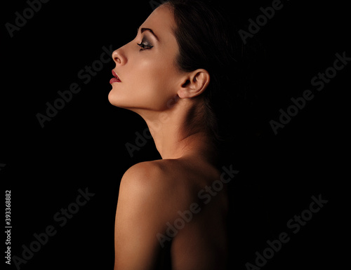 Beautiful mysterious woman in darkness with healthy neck, shoulders and serious wisdom look on dramatic black background with empty copy space. Closeup portrait. Profile view.