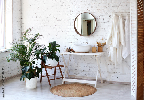 Modern bathroom interior with big round mirror, table with washbasin, big window and plants near white brick wall