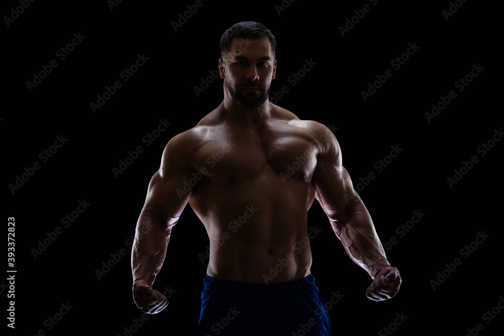Portrait of a bodybuilder standing isolated on black background in a shadow with clenched fists to show off his muscles